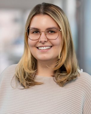 Junge Frau mit blonden Haaren und Brille, lächelt freundlich. Sie trägt ein helles, gestreiftes Oberteil und steht in einem modernen Innenraum.