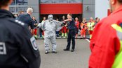 Ein Feuerwehrmann hilft einem Kollegen, einen Schutzanzug anzulegen, während eine Gruppe von Einsatzkräften zuschaut.