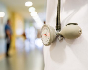 Ein Stethoskop und ein Blutdruckmessgerät hängen an der Kleidung einer Person in einem Krankenhausflur, während unscharfe Figuren im Hintergrund zu sehen sind.