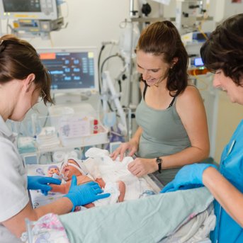 Eine Mutter hält ein Neugeborenes, während zwei Pflegekräfte bei der Versorgung des Babys helfen. Alle tragen Handschuhe.