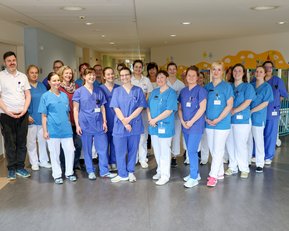 Eine Gruppe von 16 Personen in medizinischer Kleidung steht in einem Krankenhausflur, lächelnd und in zwei Reihen angeordnet.