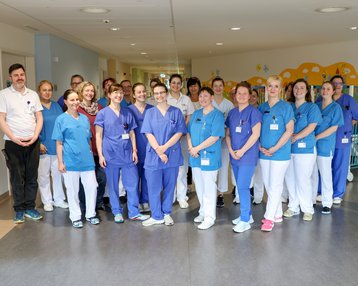 Eine Gruppe von 16 Personen in medizinischer Kleidung steht in einem Krankenhausflur, lächelnd und in zwei Reihen angeordnet.