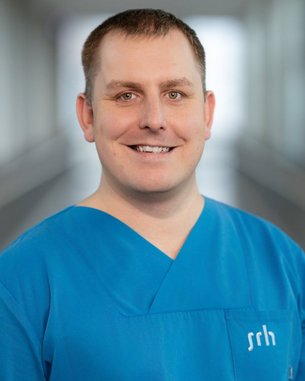 Ein Mann in blauer medizinischer Kleidung lächelt in die Kamera, mit einem neutralen Hintergrund, der eine Klinikumgebung andeutet.