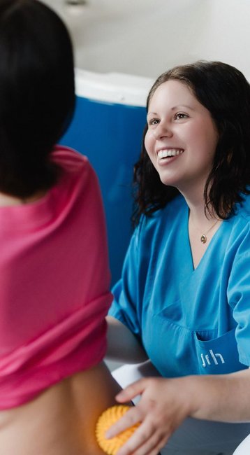 Eine schwangere Frau in einem pinken Oberteil nutzt ein Seil zur Unterstützung, während eine Fachkraft in blauer Kleidung sie anleitet.