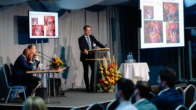 Ein Referent steht an einem Podium und präsentiert medizinische Bilder auf Bildschirmen während einer Konferenz. Im Publikum sitzen Zuhörer.
