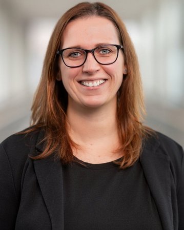 Eine lächelnde Frau mit langen, braunen Haaren und Brille trägt ein schwarzes Oberteil und einen schwarzen Blazer.