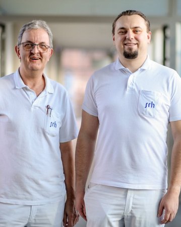 Zwei Männer in weißen Arbeitskleidung stehen in einem Flur. Beide tragen ein Namensschild mit der Aufschrift "srh".