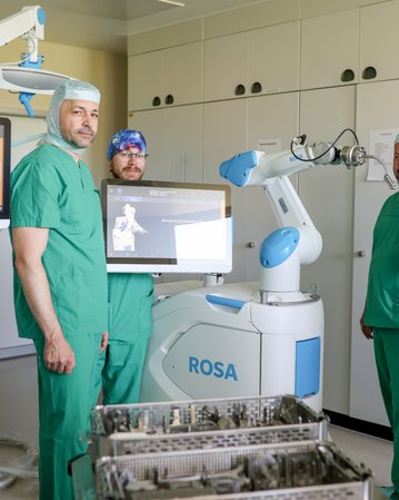 Fünf medizinische Fachkräfte in grüner OP-Kleidung stehen um einen chirurgischen Roboter namens ROSA in einem Operationssaal.