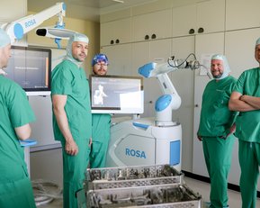 Fünf medizinische Fachkräfte in grüner OP-Kleidung stehen um einen chirurgischen Roboter namens ROSA in einem Operationssaal.