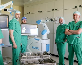 Fünf medizinische Fachkräfte in grüner OP-Kleidung stehen um einen chirurgischen Roboter namens ROSA in einem Operationssaal.