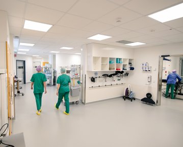 Zwei medizinische Fachkräfte in grüner Kleidung gehen durch einen modernen Krankenhausflur mit medizinischen Geräten und Regalen.