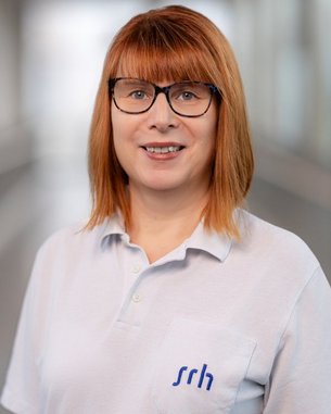 Frau mit rotem Haar und Brille trägt ein helles Poloshirt mit dem Logo "srh". Sie steht in einem modernen Innenraum.