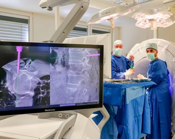 Ein Monitor zeigt medizinische Bilder während eines chirurgischen Eingriffs. Zwei Chirurgen in blauen OP-Kitteln und Masken arbeiten an einem Patienten, der sich hinter einem großen medizinischen Gerät befindet.
