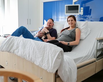 Ein Paar sitzt auf einem Krankenhausbett, während sie ein Neugeborenes halten. Beide wirken entspannt und glücklich.