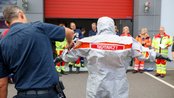 Ein Notarzt in einem Schutzanzug wird von einem Polizisten angekleidet, während eine Gruppe von Rettungskräften im Hintergrund zuschaut.