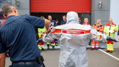 Ein Notarzt in einem Schutzanzug wird von einem Polizisten angekleidet, während eine Gruppe von Rettungskräften im Hintergrund zuschaut.