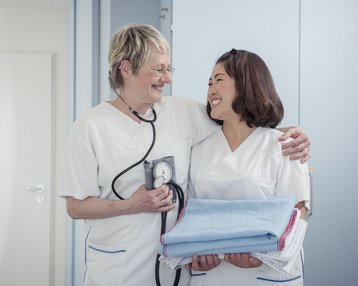Zwei Krankenschwestern stehen in einem Krankenhauszimmer, eine hält ein Stethoskop und die andere trägt einen Stapel Handtücher. Beide lächeln und scheinen sich freundlich zu unterhalten.