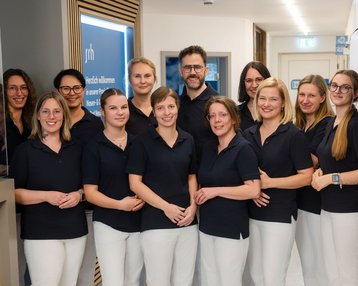 Ein Team von zwölf Personen in einheitlicher Kleidung steht in einem Flur. Sie lächeln und posieren gemeinsam für das Foto.