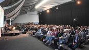 Eine große Gruppe von Menschen sitzt in einem Konferenzraum, während eine Präsentation auf einer Leinwand gezeigt wird.