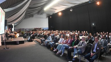Eine große Gruppe von Menschen sitzt in einem Konferenzraum, während eine Präsentation auf einer Leinwand gezeigt wird.