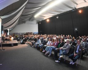 Eine große Gruppe von Menschen sitzt in einem Konferenzraum, während eine Präsentation auf einer Leinwand gezeigt wird.
