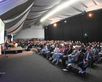 Eine große Gruppe von Menschen sitzt in einem Konferenzraum, während eine Präsentation auf einer Leinwand gezeigt wird.