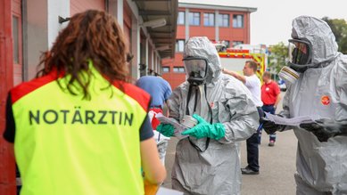 Zwei Personen in Schutzanzügen erhalten Anweisungen von einer Notärztin. Im Hintergrund sind weitere Einsatzkräfte und Fahrzeuge sichtbar.