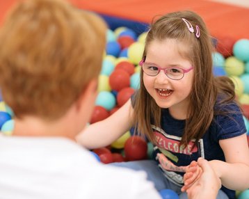 Ein fröhliches Mädchen mit Brille sitzt in einem Ballbecken und lächelt, während sie die Hände einer erwachsenen Person hält, die ihr gegenüber sitzt.
