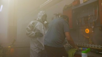 Eine Person in einem Schutzanzug steht neben einer anderen Person, die an einem Tisch arbeitet, umgeben von Dampf oder Rauch.