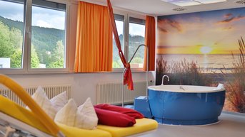 Ein modernes Badezimmer mit einer blauen Badewanne, einem gelben Liegestuhl und roten Kissen. Große Fenster mit orangefarbenen Vorhängen bieten einen Blick auf die Natur. An der Wand ist ein großes Bild eines Sonnenuntergangs am Strand.