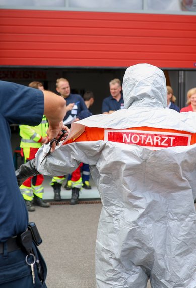 Ein Notarzt in Schutzanzug wird von einem Feuerwehrmann angeleitet, während eine Gruppe von Rettungskräften zuschaut.