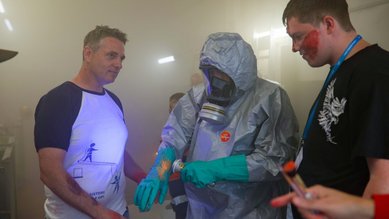Drei Personen in einem rauchigen Raum, eine Person trägt einen Schutzanzug und Handschuhe, während die anderen zuschauen.