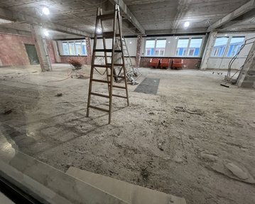 Leiter steht in einem unvollendeten Raum mit freiliegendem Boden und Fenstern im Hintergrund. Baustellenmaterialien sind sichtbar.