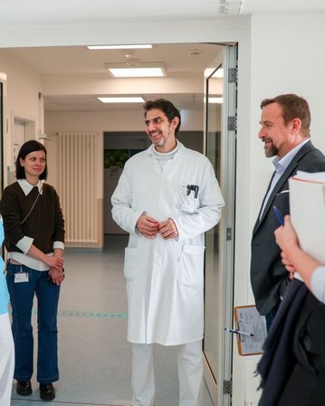 Eine Gruppe von sechs Personen, darunter Ärzte und Pflegekräfte, steht in einem Krankenhausflur und führt ein Gespräch.