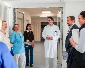 Eine Gruppe von sechs Personen, darunter Ärzte und Pflegekräfte, steht in einem Krankenhausflur und führt ein Gespräch.