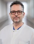 Ein Mann mit Brille und Bart trägt einen weißen Kittel. Er steht in einem hellen Flur und schaut direkt in die Kamera.