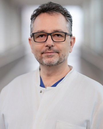 Ein Mann mit Brille und Bart trägt einen weißen Kittel. Er steht in einem hellen Flur und schaut direkt in die Kamera.