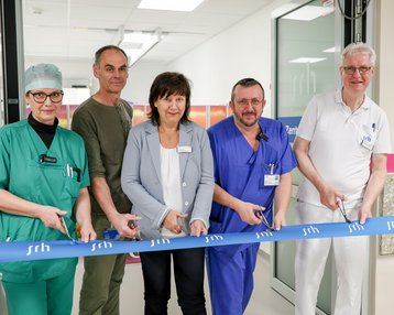 Fünf Personen in Arbeitskleidung schneiden ein blaues Band durch, um eine Eröffnung in einer medizinischen Einrichtung zu feiern.