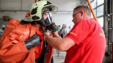 Eine Person in einem orangefarbenen Schutzanzug wird von einem Ausbilder mit einer Atemmaske ausgestattet. Im Hintergrund sind weitere Personen zu sehen.