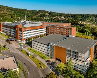 Moderne Gebäude mit orangefarbenen Fassaden und großen Fenstern, umgeben von Grünflächen und Bäumen. Die Architektur zeigt eine Kombination aus mehreren Baukörpern, die sich harmonisch in die Landschaft einfügen.