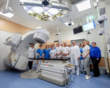 Ein Team von medizinischem Personal steht in einem modernen Behandlungsraum mit einem Bestrahlungsgerät und technischen Geräten.