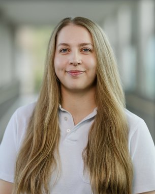 Eine Person mit langen, blonden Haaren trägt ein weißes Polohemd und steht in einem hellen, modernen Innenraum.