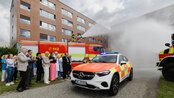 Ein Einsatzfahrzeug der Feuerwehr steht vor einem Gebäude, während Feuerwehrleute Wasser spritzen. Zuschauer applaudieren im Hintergrund.