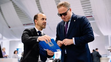 Zwei Männer in Anzügen diskutieren über ein blaues Dokument. Einer trägt Sonnenbrillen, während der andere auf das Dokument zeigt.