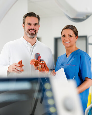 Ein Arzt und eine Krankenschwester stehen in einem modernen medizinischen Raum. Der Arzt hält ein anatomisches Modell eines Herzens, während die Krankenschwester ein Dokument in der Hand hält. Im Hintergrund sind medizinische Geräte und Bildschirme sichtbar.