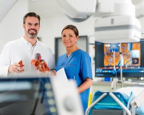 Ein Arzt und eine Krankenschwester stehen in einem modernen medizinischen Raum. Der Arzt hält ein anatomisches Modell eines Herzens, während die Krankenschwester ein Dokument in der Hand hält. Im Hintergrund sind medizinische Geräte und Bildschirme sichtbar.