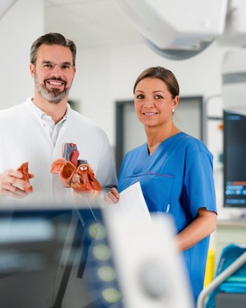 Ein Arzt und eine Krankenschwester stehen in einem modernen medizinischen Raum. Der Arzt hält ein anatomisches Modell eines Herzens, während die Krankenschwester ein Dokument in der Hand hält. Im Hintergrund sind medizinische Geräte und Bildschirme sichtbar.